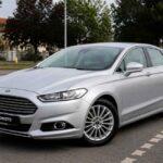 Srebrny Ford Mondeo zaparkowany na brukowanym parkingu przy ulicy drzewa i zabudowania w tle