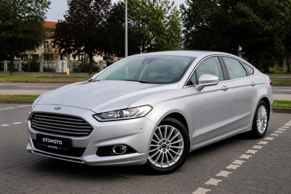 Srebrny Ford Mondeo zaparkowany na brukowanym parkingu przy ulicy drzewa i zabudowania w tle