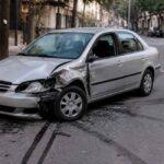 Srebrny sedan z uszkodzonym przodem i bokiem stoi na miejskiej ulicy po kolizji