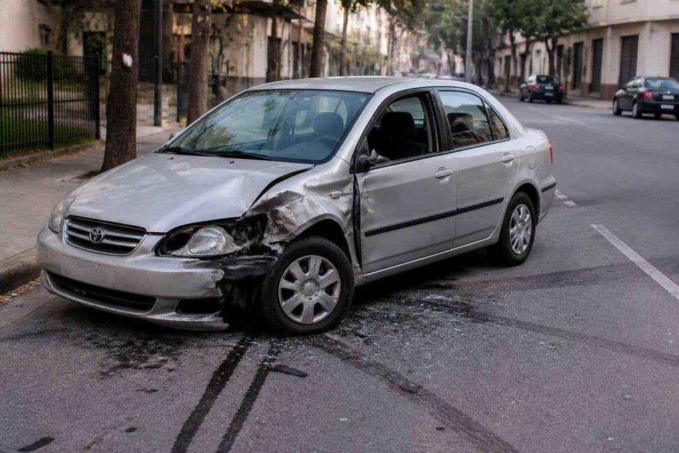 Srebrny sedan z uszkodzonym przodem i bokiem stoi na miejskiej ulicy po kolizji